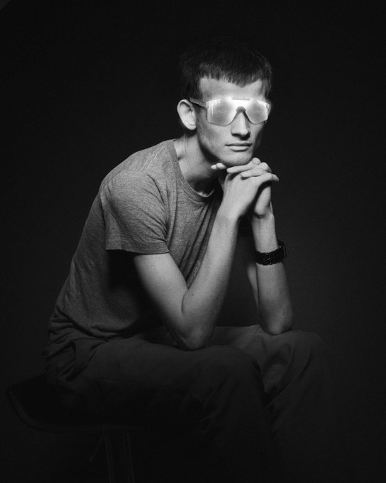 A black-and-white portrait of a young man seated against a dark background, wearing reflective sunglasses, a gray t-shirt, and a watch, with his hands clasped under his chin.