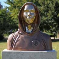 A bronze bust sculpture depicting a hooded figure with a reflective gold face, mounted on a stone pedestal outdoors.