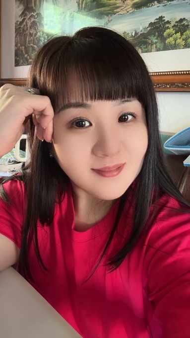 A selfie of a young woman with dark hair and bangs wearing a red t-shirt, with a framed landscape painting visible on the wall behind her.