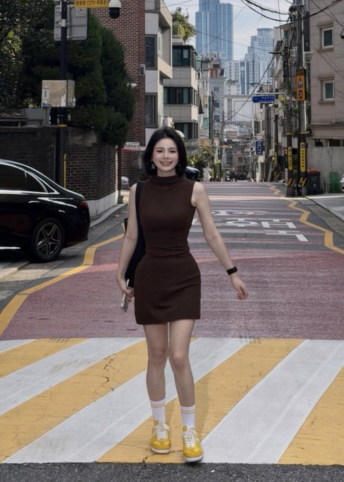 A young woman in a brown sleeveless mini dress, white socks, and yellow sneakers stands on a yellow-and-white crosswalk in a city street.
