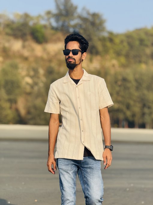 A bearded man wearing sunglasses, a cream-colored short-sleeve striped shirt, and jeans stands outdoors with a blurred hillside in the background.