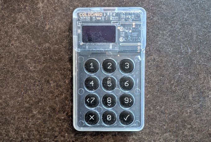 A transparent, plastic Coldcard Mk4 hardware cryptocurrency wallet device with a numeric keypad resting on a textured surface.