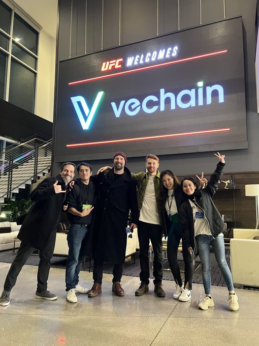A group of seven people posing in front of a UFC Welcomes VeChain digital screen display.