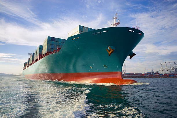 A large teal and red container ship sails through choppy blue waters, with a port visible in the distance under a partly cloudy sky.