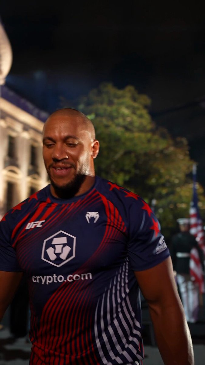 The image features a man wearing an orange, blue, and red shirt with white text that says " UFC White Houe" printed on it. The man appears to be walking down a street at night, possibly attending a special event or gathering related to the UFC.