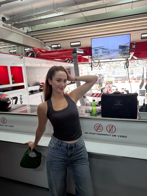 A woman poses playfully at what appears to be an F1 race paddock garage, with a "NO PHOTOGRAPHY OR VIDEO" sign visible on the barrier and "47" markings on equipment behind her. The "race day" caption fits perfectly as she's clearly enjoying the F1 atmosphere, with helmets and racing gear visible in the background alongside a track-view monitor.