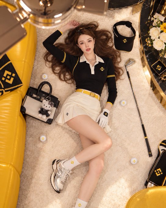 A woman with long brown hair lies on a carpet wearing a black and yellow polo shirt and white skirt, surrounded by luxury accessories including golf clubs and a designer bag.