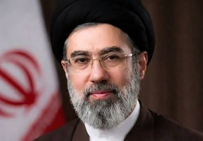 Portrait of an Iranian cleric with a black turban, glasses, and a gray beard, posed in front of an Iranian flag.