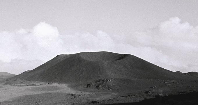 Black and white photograph of a barren, mountainous landscape with a dark, conical hill or cinder cone in the foreground under a cloudy sky.