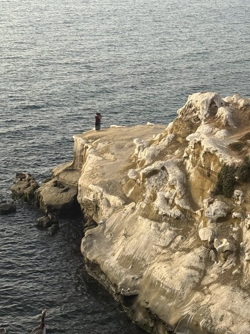 A person standing at the edge of a tall sandstone cliff overlooking the ocean, with rugged rock formations and dark blue water below.
