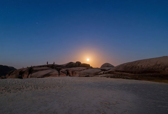 A nighttime photograph of a rocky coastal landscape with a bright moon on the horizon, stars in the sky, and small figures standing on the boulders.
