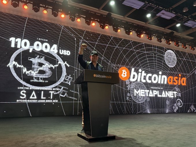 A man in a black shirt and cap standing at a podium at a Bitcoin Asia conference, holding up a phone, with a large screen showing "110,004 USD" and logos for bitcoin asia, SALT, and METAPLANET.