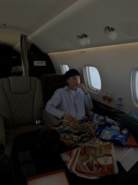 A young man wearing a blue shirt sits inside the cabin of a private jet, surrounded by an assortment of packaged snacks spread across the seat and tray table.