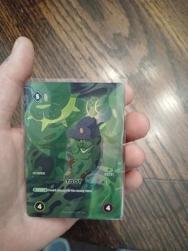 A hand holds a Pokémon trading card featuring the character Toedscruel, a green, cactus-like creature with multiple limbs, against a wooden floor background.