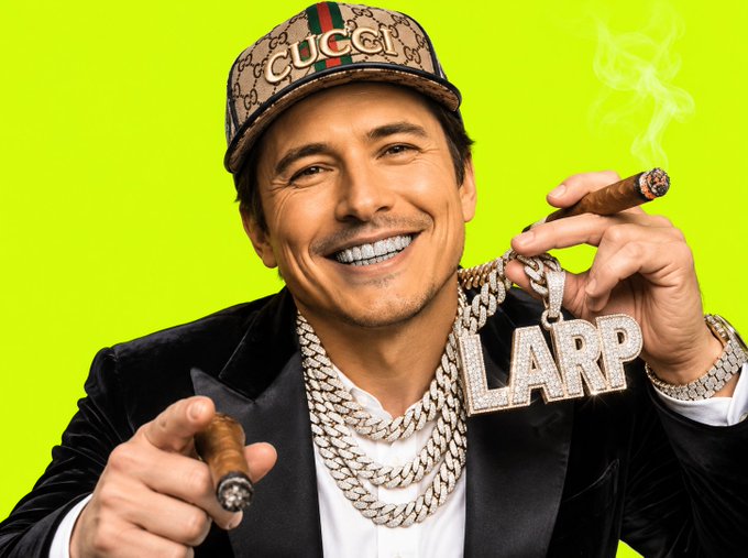 A man smiling brightly while holding a diamond-encrusted 'LARP' pendant and two cigars, wearing a Gucci cap, multiple chains, and a black blazer against a neon green background.