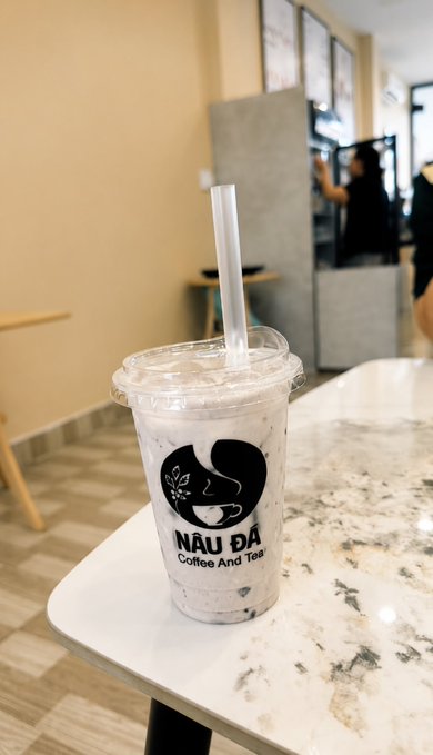 A plastic cup of iced Thai tea from 'NẤU ĐÁ Coffee And Tea' with a white straw, sitting on a marble table in what appears to be a coffee shop.