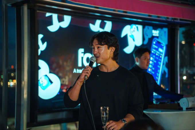 A man in a black sweater speaks into a microphone while holding a champagne flute at what appears to be an evening event, with a large illuminated display featuring stylized characters and a "fc" logo behind him, while another man in dark attire stands nearby—fitting the "KBW signoff" caption as a closing moment at the Korea Blockchain Week conference.