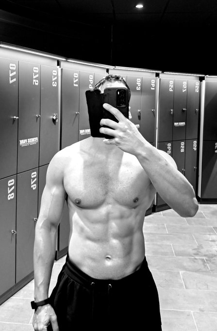 A black-and-white photo shows a shirtless, muscular man taking a mirror selfie in a locker room with numbered lockers in the background.