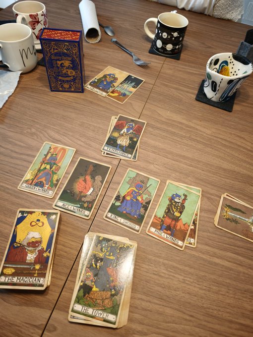 A tarot card reading setup on a wooden table featuring cards like "THE MAGICIAN", "THE TOWER", "THE FOOL KNIGHT", and "THE BATON KING" alongside several coffee mugs.