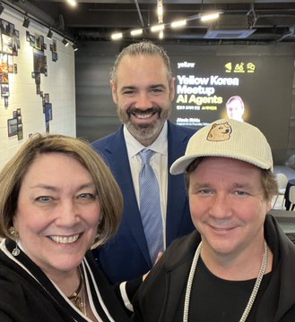Three people pose for a photo at what appears to be a Yellow Korner Meetup, with a screen in the background reading 'Yellow Korner Meetup Art Agents' and 'Yellow Korner'.