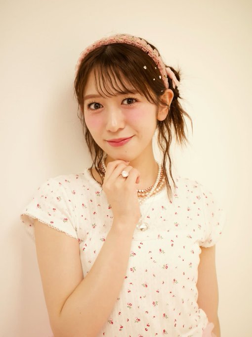 A young woman with brown hair styled in a side ponytail, wearing a pink crocheted headband with a bow, poses with her hand near her chin while wearing a floral blouse and pearl necklace. The soft, cheerful portrait complements the bright "Good morning" greeting and signals an upbeat start to the tour's "first day" with her "ALT" prediction.
