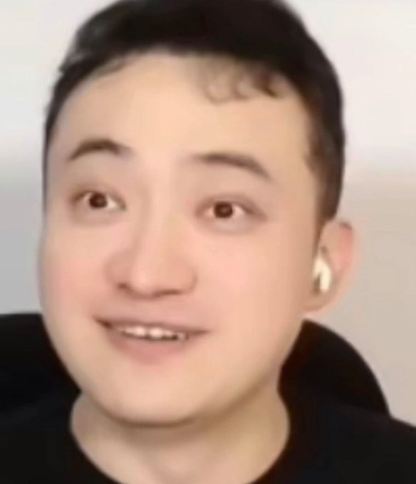 A close-up of a young man with short black hair wearing a black shirt and white wireless earbuds, smiling slightly at the camera.