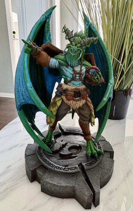 A detailed figurine of a green dragon-like creature with large wings, holding a dark orb, standing on a circular stone base.
