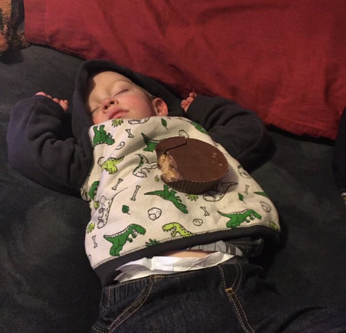 A baby sleeping on a dark blanket while wearing a dinosaur-patterned bib, with a partially eaten chocolate peanut butter cup resting on their chest.