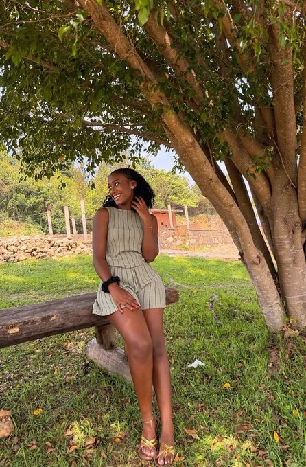 A woman in a striped dress sits on a wooden bench under a tree in a grassy outdoor setting.