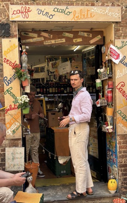 A man in a pink striped shirt and white pants stands in the doorway of an Italian shop with a painted sign advertising 'caffè, vino, aceto, birra, zucchero' and other goods, with another man visible inside near shelves of bottles.