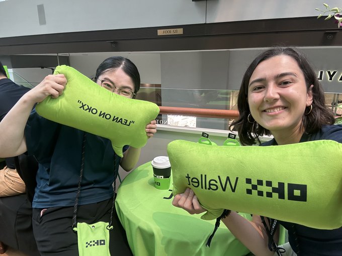 Two smiling women at a DXO Wallet booth hold up bright green bone-shaped pillows, one reading 'LEAVIN ON OKX' and the other 'OKX Wallet'.