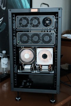 A custom-built cryptocurrency mining rig in a black open-air frame with multiple cooling fans, power supplies, and GPUs visible.