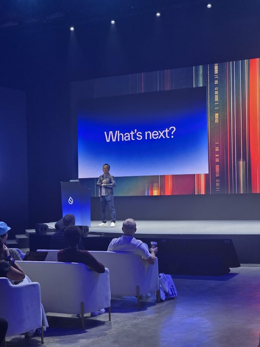 A man standing on a stage in front of a large screen displaying 'What's next?', giving a presentation to an audience seated in white armchairs.