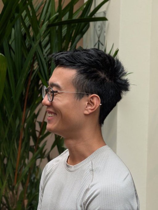 A side profile shot of a smiling young man with dark hair and glasses, wearing a grey long-sleeved shirt, standing outdoors in front of green plants.