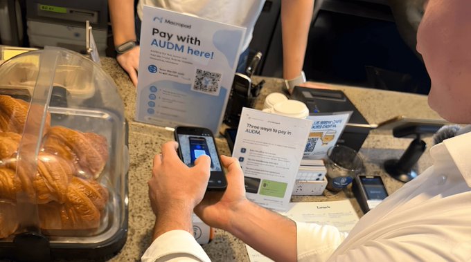 The image features two individuals at a café paying for their orders using cell phones. One person is holding up their phone displaying an electronic payment system like Audubon, while the other person is behind them writing down the order details. The scene takes place in a modern café with comfortable furnishings and a counter where the customers can make payments. There are also some pastries on display at the front of the counter for sale or ordering.