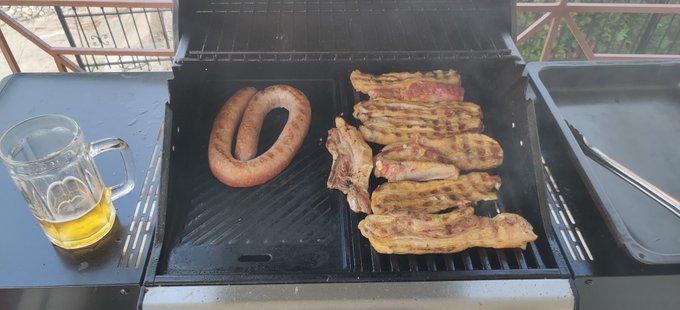 A barbecue grill cooking meat including a coiled sausage, pork belly slices, and what appears to be steak, with a glass beer mug sitting on the side table.