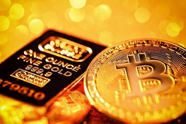 A close-up image of a Bitcoin coin next to a one-ounce gold bar (999.9 fine gold) with a golden bokeh background.