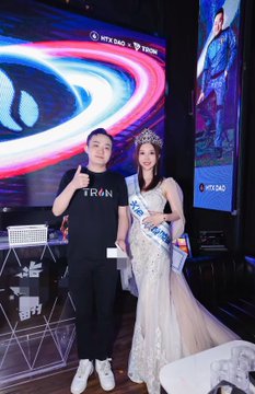 A woman wearing a "TRON" shirt stands next to a woman in a sparkly gown and tiara with a "TRON" sash, posing in front of a screen displaying "HTX DAO" and "TRON" branding. The image relates to the post by commemorating the anniversary of joining Huobi, with the pageant-style appearance connecting to the mention of winning the "Huobi Top Sister" championship.