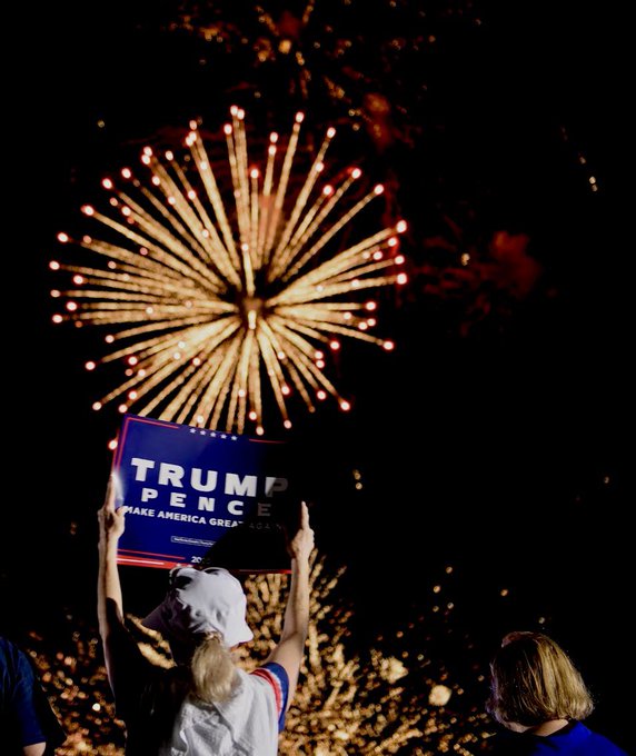 A person holding up a 'TRUMP PENCE MAKE AMERICA GREAT AGAIN' campaign sign while watching a fireworks display light up the night sky.