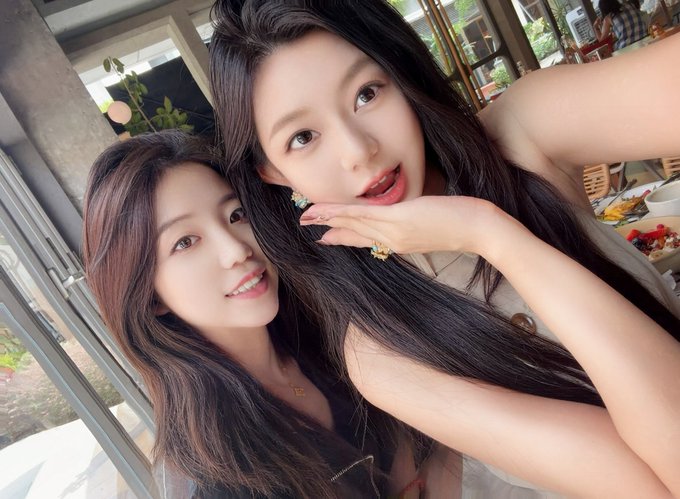 A selfie of two young women with long dark hair posing closely together at a restaurant table, with a plate of food visible in the background.