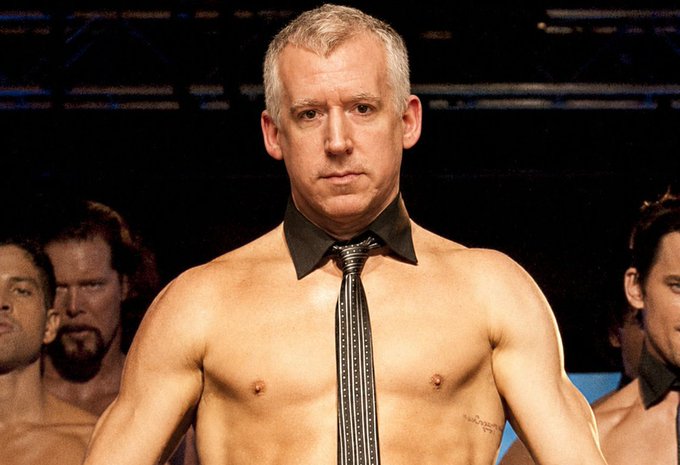 A shirtless muscular man with gray hair wears only a black shirt collar and a black-and-silver striped tie, standing in a wrestling ring with other men visible in the background.