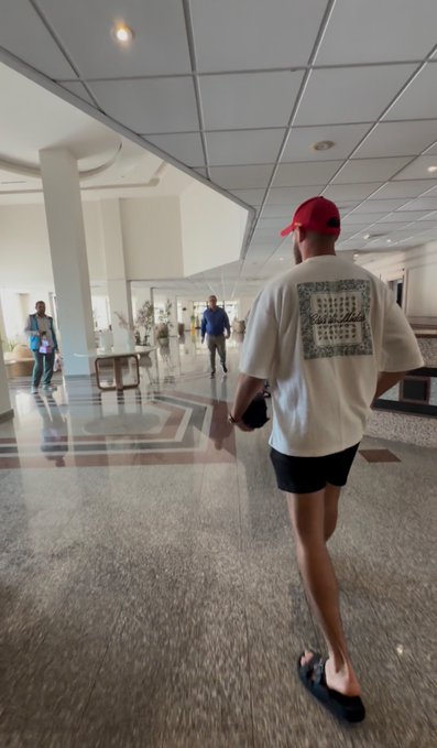 A person wearing a red cap and white graphic t-shirt walks through a bright, modern hotel lobby with polished marble floors. The casual, well-lit setting complements the cheerful "Gm friends" greeting, suggesting a relaxed morning moment.