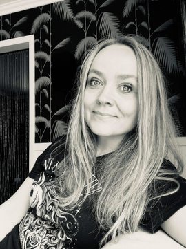 A black-and-white selfie of a woman with long blonde hair wearing a black graphic t-shirt, posing in front of dark tropical leaf wallpaper.