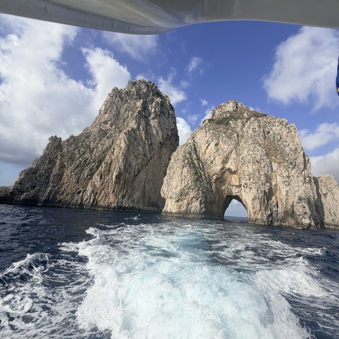 A boat cruises past the iconic arch rock formation of Cabo San Lucas under a partly cloudy sky. The massive natural stone gateway rising from the turquoise sea echoes the "GM" greeting as a bold "good morning" salute from the open water.
