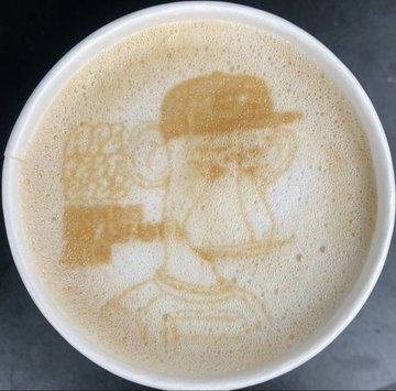 A cup of coffee with latte art depicting a cartoon character wearing a baseball cap.