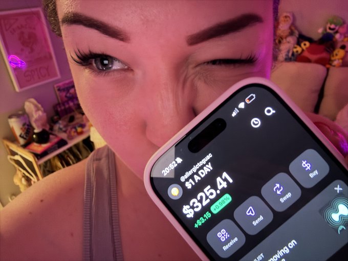 A woman holding up a phone displaying a crypto app screen showing '$1A DAY' and '$325.41' total with a +$3.15 (0.95%) gain.