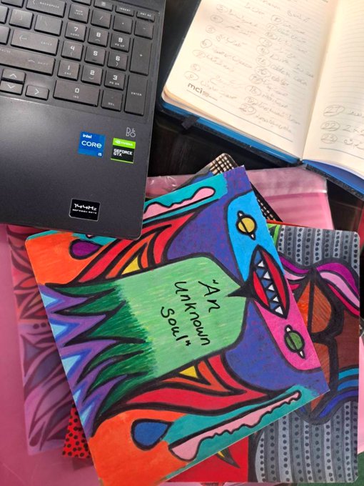 A workspace featuring a black Lenovo laptop with Intel Core i5 and NVIDIA GeForce RTX stickers, next to a colorful abstract painting with the handwritten text 'Art Unfamous Society'.
