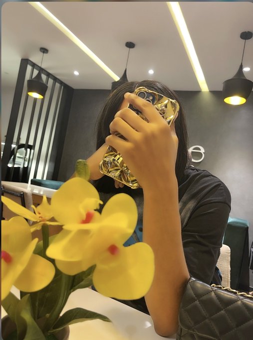 A mirror selfie-style photo showing a person holding a gold-textured smartphone in front of their face, with a bouquet of yellow orchids in the foreground and a black quilted handbag visible.