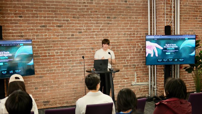 A presenter stands at a laptop in front of a brick wall, with two screens displaying a presentation titled 'Build & Trade the Next Agentic Algorithm', while an audience watches.