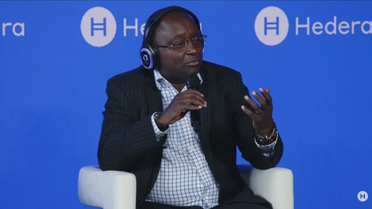 The image features a man wearing headphones, sitting in a chair, and talking into a microphone. He appears to be engaged in a conversation or presentation related to Kenya's economy and sustainability efforts. The background of the image shows a blue wall with white text that reads "@hedera" and "HEDERA". This suggests that the man is speaking at an event organized by @hedera, possibly discussing how they are helping bring Kenya to the future while addressing climate change challenges.